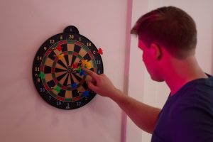 Darts: Mann deutet auf Dartsscheibe mit bunten Pfeilen